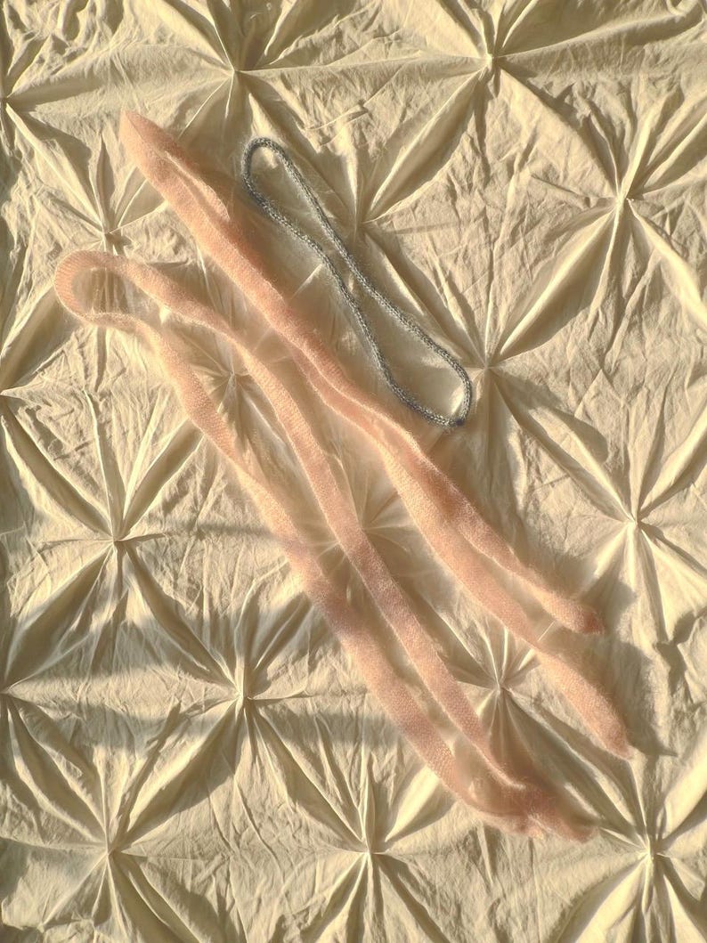 May include: Two pairs of light pink, fuzzy leg warmers and a silver beaded necklace are arranged on a cream-colored, textured surface. The leg warmers are long and slender, with a soft, textured appearance.