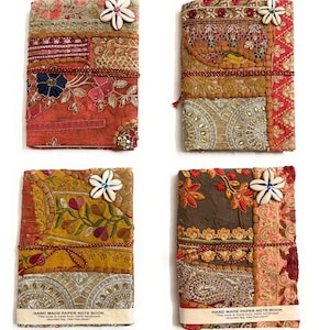 Könnte beinhalten: Vier rechteckige Notizbücher mit Patchwork-Stoffeinbänden in verschiedenen Farben wie Rot, Gold und Braun. Jeder Einband ist mit aufwendigen Stickereien, Metallic-Fäden und einer weißen Blume aus Kaurimuscheln verziert. Eine rote Kordel umschließt jedes.