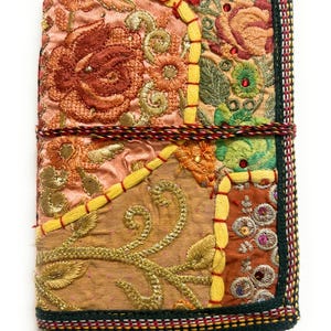 Könnte beinhalten: Ein rechteckiger Notizbuchumband aus einem Patchwork bestickter Stoffe in Pfirsich-, Gold-, Orange- und Grüntönen. Die Oberfläche ist mit Blumenmustern, metallischem Goldfaden, kleinen Perlen und Pailletten verziert und mit einer Kordel verschlossen.