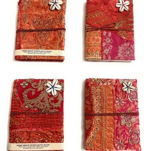 May include: Four rectangular notebooks with fabric covers in shades of orange, red, and pink, featuring gold embroidery, sequins, and a white cowrie shell accent. Each notebook is secured with a thin dark cord and includes a paper band with text.