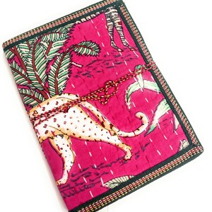 May include: A hot pink journal featuring a cheetah print and tropical leaf design. It is bound with a red and white cord. The edges are trimmed with a dark green and gold border.