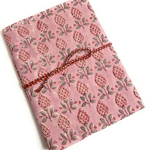 Może przedstawiać: Złożony, prostokątny przedmiot z tkaniny z powtarzającym się wzorem czerwonych i białych kwiatowych wzorów na różowym tle. Czerwony, żółty i czarny skręcony sznurek owija przedmiot. Tkanina wydaje się być bawełną lub podobnym materiałem.