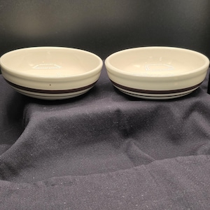 May include: Two round, cream-colored ceramic bowls featuring a dark brown horizontal stripe around the exterior. The interior of each bowl is smooth and light-colored, with a simple, clean design suitable for serving food or holding small items.