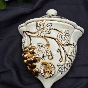 May include: White ceramic wall-mounted wine dispenser with a lid. The dispenser is decorated with gold-colored grape clusters and vines. The dispenser has a gold-colored spigot and a small, round lid handle.