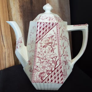 May include: Cream-colored ceramic teapot with a red floral pattern and geometric accents. The teapot has a lid with a small knob, a spout, and a handle. The design features flowers and foliage, creating a vintage aesthetic.