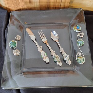 May include: A clear square glass plate with a knife, fork, and spoon. The utensils have silver handles with wire and green bead accents. The plate has decorative wire and green bead accents on the corners.