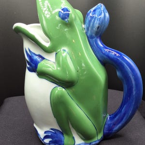 May include: A ceramic pitcher in the shape of a frog. The frog is green with blue accents on the eyes, feet, and handle. The pitcher's body is white. The handle is a curved blue shape. The frog is in a sitting position, with its mouth open.