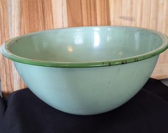 Puede incluir: Un cuenco de esmalte verde claro con un borde verde oscuro. El cuenco tiene forma redondeada y está sobre un fondo de madera. El cuenco es probablemente vintage y muestra signos de desgaste.