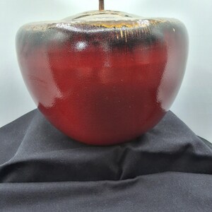 May include: A red ceramic bowl with a lid and a bronze handle. The bowl has a glossy finish and a dark rim. The handle is a curved, horizontal bar. The bowl is sitting on a black surface.