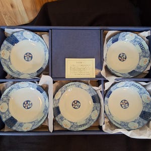 May include: A set of six ceramic bowls with a blue and white floral design, presented in a blue box. Each bowl features a central pattern and a patterned border. The box contains a small card with Japanese text.