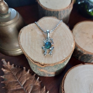 Puede incluir: Un collar con un colgante de rana de plata con una incrustación de concha de abulón verde. El collar está sobre una superficie de madera.