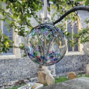 May include: A clear glass bauble with internal floral designs, suspended by a white ribbon. The bauble reflects the surrounding greenery and a stone building. The colours include blue, purple, green, and red. The bauble is approximately 8 cm in diameter.