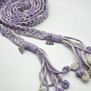 May include: A long, braided cord with a purple and gray color scheme. The cord has two tassels with knotted ends and silver Celtic knot charms.