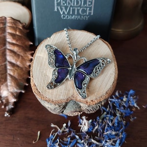 Puede incluir: Un collar de plata con un colgante de mariposa de color púrpura iridiscente. El collar está sobre una superficie de madera con pétalos de flores azules esparcidos a su alrededor.