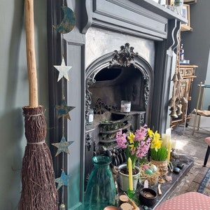 Puede incluir: Una repisa de chimenea decorada con una guirnalda de estrellas y medias lunas. La chimenea es negra y tiene un hogar de azulejos blancos. Hay velas y flores en la repisa, y una escoba apoyada contra la pared.