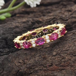 Peut inclure: Une bague en or ornée d'une rangée continue de pierres précieuses roses foncées de forme ovale, entrecoupées de petites pierres claires rondes. La bague est posée sur un fond sombre et texturé, mettant en valeur l'éclat des gemmes.