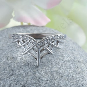 May include: A silver-colored ring with a unique design. The ring features a V-shaped band with small, clear stones and an intricate, web-like pattern below the band. The ring is set on a gray stone.