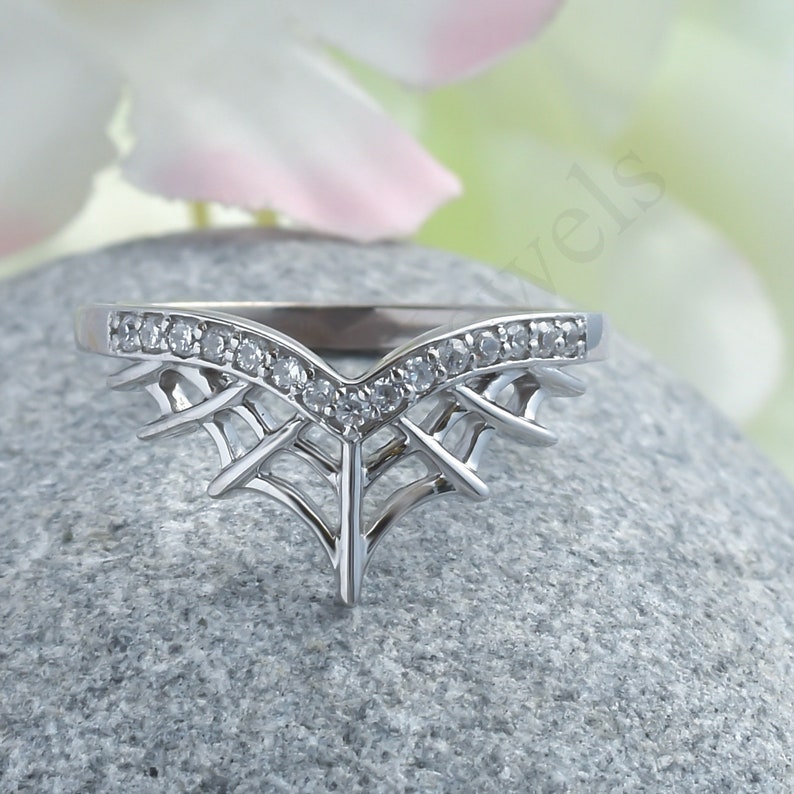 May include: A silver-colored ring with a unique design. The ring features a V-shaped band with small, clear stones and an intricate, web-like pattern below the band. The ring is set on a gray stone.