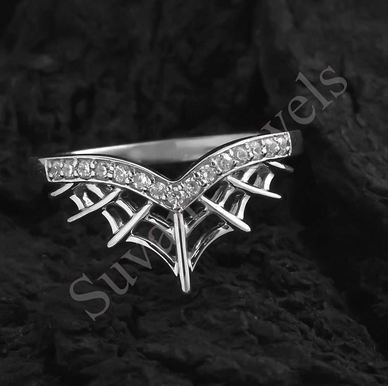 May include: A silver-colored ring with a V-shaped design, adorned with small, clear gemstones along the top curve. The ring features a unique, web-like pattern beneath the gemstones, adding a distinctive flair. The ring is set against a dark, textured background.