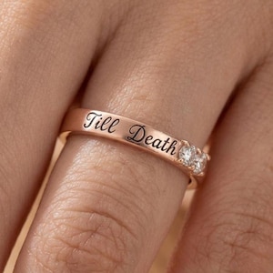 Può includere: Un anello a fascia in oro rosa con il testo Till Death inciso in nero sulla superficie esterna. L'anello è impreziosito da due piccole pietre trasparenti a taglio rotondo, posizionate una accanto all'altra vicino all'incisione sul metallo.