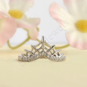 May include: A silver-toned tiara-shaped ring with a row of small, clear gemstones along the base. The ring has an openwork design with a crown-like shape. The background features soft pink and yellow tones.