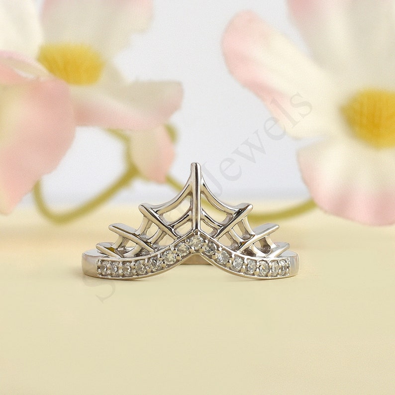 May include: A silver-toned tiara-shaped ring with a row of small, clear gemstones along the base. The ring has an openwork design with a crown-like shape. The background features soft pink and yellow tones.