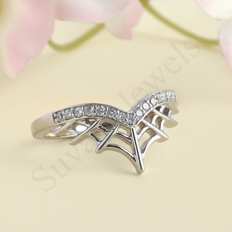 May include: A silver-colored ring with a unique spiderweb design. The ring features a V-shaped band with a row of small, clear stones along the top. The design includes a delicate web-like pattern on the sides.
