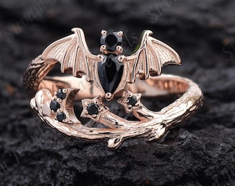 May include: Rose gold ring featuring a bat with black gemstone accents. The bat has outstretched wings and is centered above a branch with crescent moons and star-shaped gemstones. The ring is set against a dark, textured background.