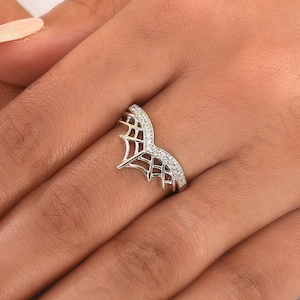 May include: A silver ring with a spiderweb design. The ring features a curved band with a row of small, clear gemstones. The spiderweb design is on the front of the ring, adding a unique and delicate touch.