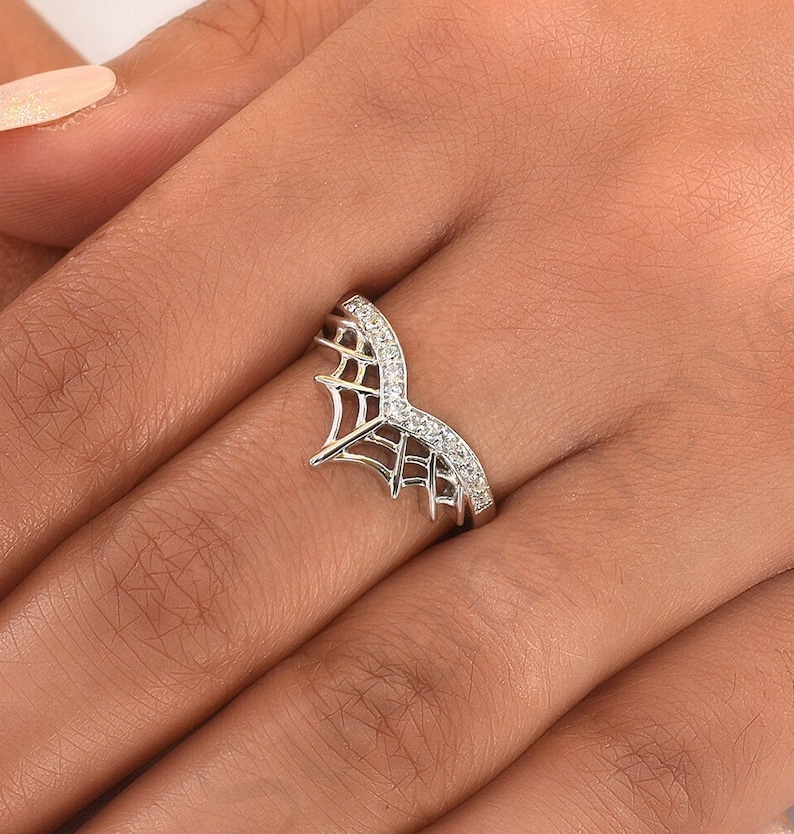 May include: A silver ring with a spiderweb design. The ring features a curved band with a row of small, clear gemstones. The spiderweb design is on the front of the ring, adding a unique and delicate touch.