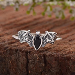 Può includere: Un anello in argento con un elemento centrale a forma di pipistrello con ali spiegate e una gemma nera a goccia al centro. La superficie metallica presenta incisioni dettagliate sulle ali, in contrasto con la pietra scura e liscia.