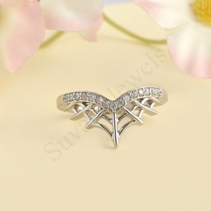 May include: A silver-toned ring with a V-shaped design, adorned with small, clear gemstones along the top curve. The ring features an open, web-like pattern beneath the gemstones, creating a delicate and intricate look. The ring is set against a soft, neutral background.