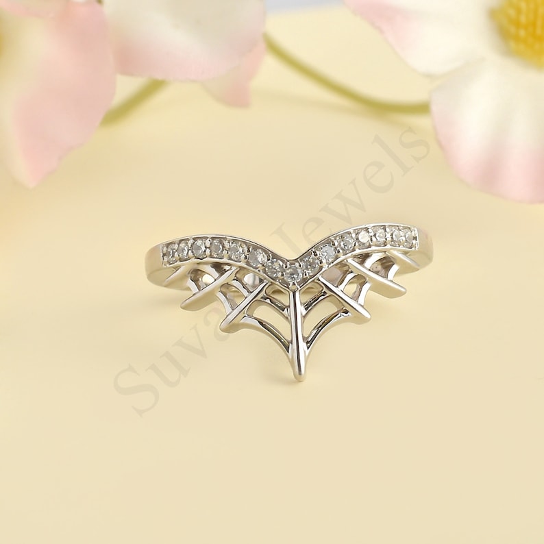 May include: A silver-toned ring with a V-shaped design, adorned with small, clear gemstones along the top curve. The ring features an open, web-like pattern beneath the gemstones, creating a delicate and intricate look. The ring is set against a soft, neutral background.