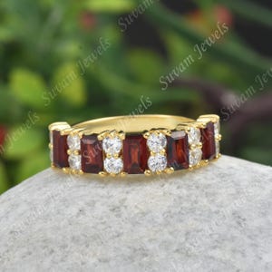 May include: A gold ring with alternating rectangular red and round white gemstones. The ring is displayed on a light grey surface with a blurred green background.