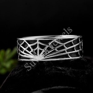 Könnte beinhalten: Ein silberner Ring mit einem detaillierten Spinnennetz-Design. Der Ring hat einen dunklen Hintergrund, der mit dem hellen silbernen Netzmuster kontrastiert. Der Ring ist im Bandstil gehalten, wobei sich das Netzdesign um den gesamten Umfang wickelt.