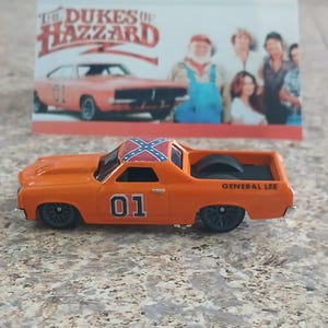 May include: An orange toy car with the number "01" on the side and a Confederate flag on the roof. The words "GENERAL LEE" are printed on the back. A card in the background features the text "The Dukes of Hazzard" and images of the show's cast and car.