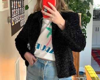 May include: A black textured jacket with a zipper closure is worn over a white t-shirt with a palm tree graphic and the word "JUBA". The person is wearing light blue jeans. A red phone case is visible.