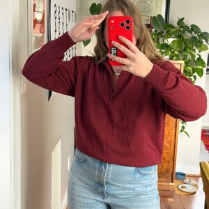 Puede incluir: Un su&eacute;ter burdeos de manga larga con cuello con cremallera. El su&eacute;ter se combina con vaqueros de mezclilla azul claro. La persona de la imagen sostiene un tel&eacute;fono rojo.