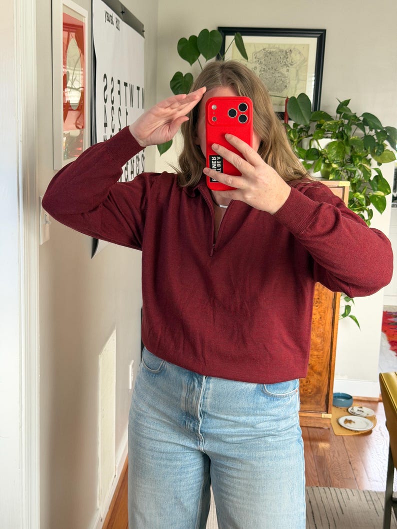 Puede incluir: Un su&eacute;ter burdeos de manga larga con cuello con cremallera. El su&eacute;ter se combina con vaqueros de mezclilla azul claro. La persona de la imagen sostiene un tel&eacute;fono rojo.
