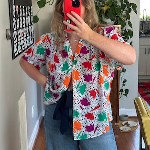May include: A short-sleeved button-up shirt with a white background and a colorful floral pattern in orange, red, green, and purple. The shirt is paired with blue jeans. A navy blue item is tied around the waist.