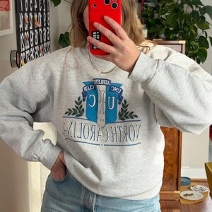 May include: A light gray sweatshirt with the words "NORTH CAROLINA" in blue and white, and a blue and white emblem. The sweatshirt has long sleeves and a crew neck. The person in the image is wearing blue jeans.
