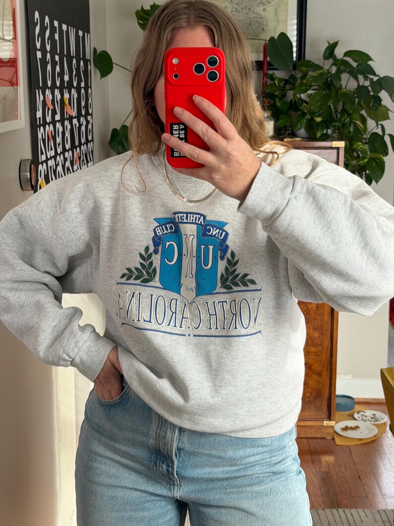 May include: A light gray sweatshirt with the words "NORTH CAROLINA" in blue and white, and a blue and white emblem. The sweatshirt has long sleeves and a crew neck. The person in the image is wearing blue jeans.