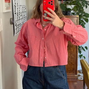 Könnte beinhalten: Ein gecropptes Hemd mit rot-weißem Streifenmuster und langen Ärmeln, geknöpft. Das Hemd wird mit einer dunkelblauen Jeans getragen. Das Outfit ist lässig und für den Alltag geeignet.
