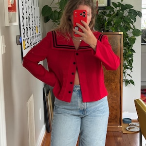 May include: A bright red long-sleeve jacket featuring a sailor-style collar with two black stripes, a vertical pleated front panel, and a row of five black buttons. The jacket is paired with light blue denim jeans with a high-waisted fit.