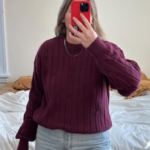 May include: A burgundy cable knit sweater with a small embroidered logo on the front. The sweater is worn over light blue jeans.