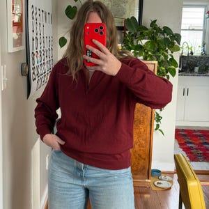Puede incluir: Un su&eacute;ter burdeos de manga larga con cuello con cremallera. El su&eacute;ter se combina con vaqueros azules de lavado claro. La persona sostiene un tel&eacute;fono rojo frente a su rostro. Un gran p&oacute;ster con texto negro es visible en la pared.