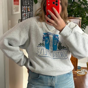 May include: A light gray sweatshirt with the text "UNC ATHLETIC CLUB NORTH CAROLINA" in blue and white. The sweatshirt is paired with light blue jeans.