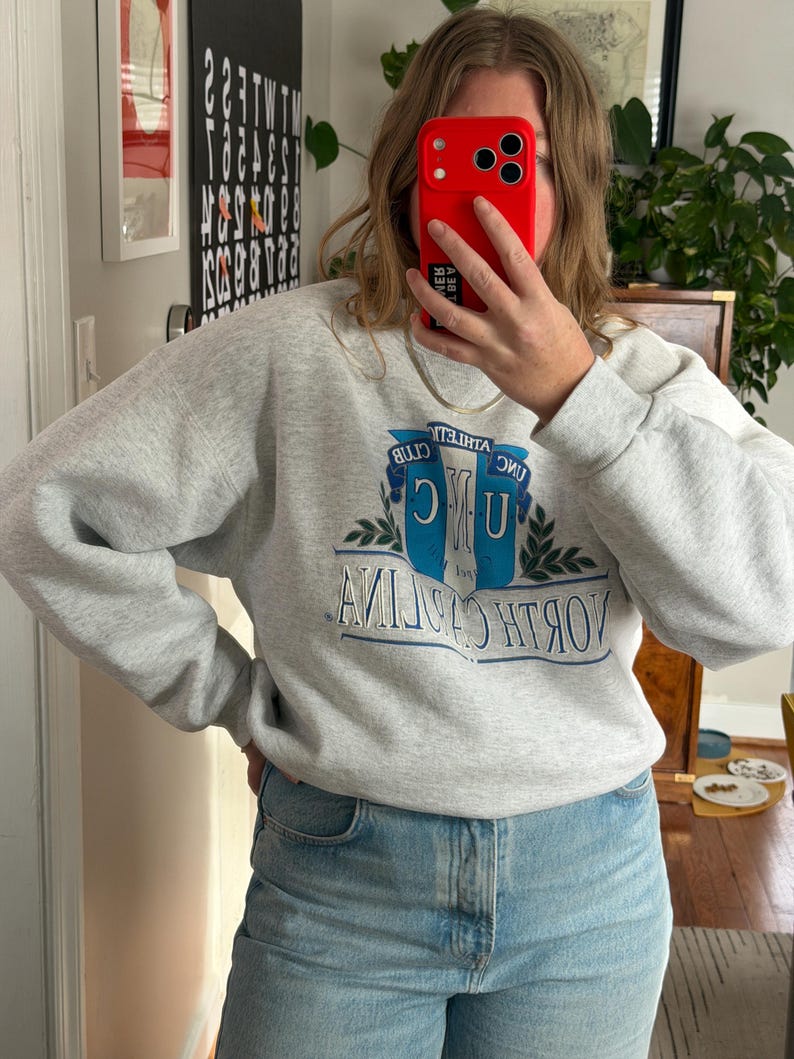 May include: A light gray sweatshirt with the text "UNC ATHLETIC CLUB NORTH CAROLINA" in blue and white. The sweatshirt is paired with light blue jeans.