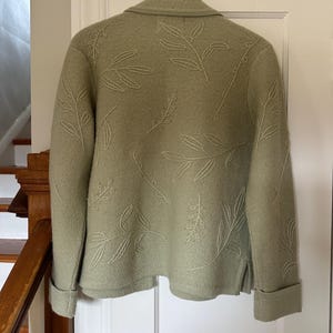 May include: Sage green jacket with embroidered leaf design. The jacket features a collared neckline, long sleeves with folded cuffs, and a button closure. The leaf pattern is repeated across the back and sleeves. The jacket is hanging on a wooden hanger.