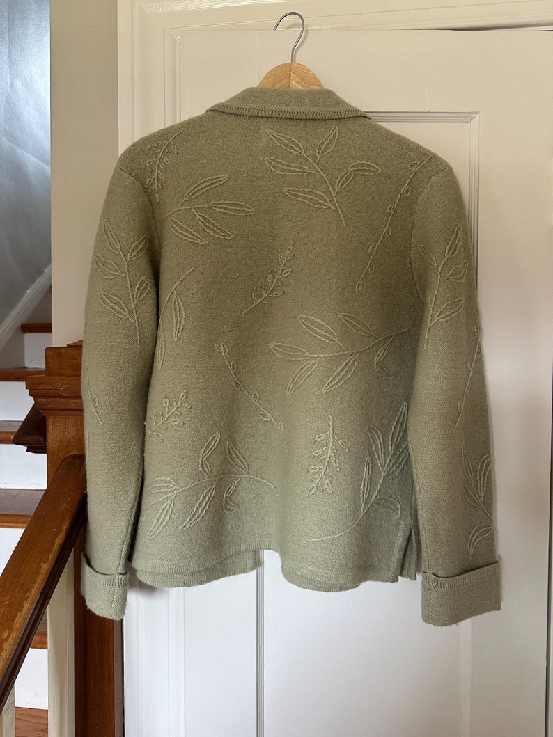 May include: Sage green jacket with embroidered leaf design. The jacket features a collared neckline, long sleeves with folded cuffs, and a button closure. The leaf pattern is repeated across the back and sleeves. The jacket is hanging on a wooden hanger.
